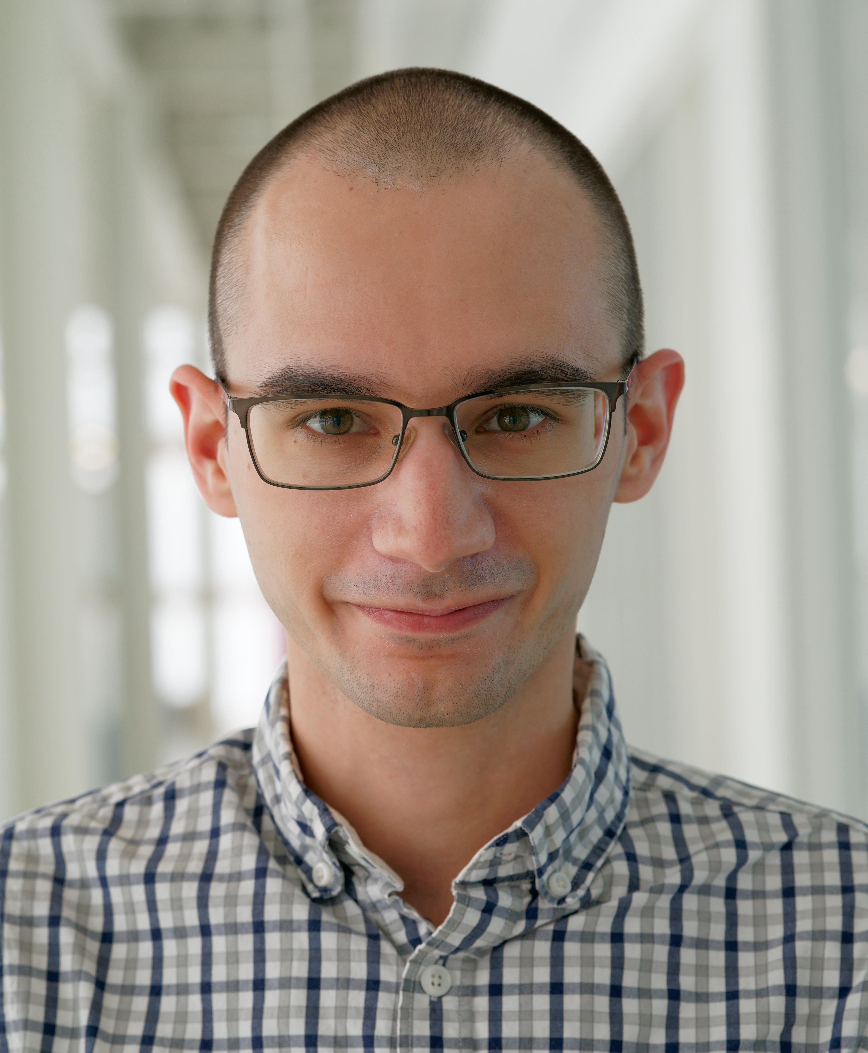 A headshot of Noah, looking into the camera and smiling slightly. He has a shaved head and glasses, and is wearing a striped button-down shirt.
