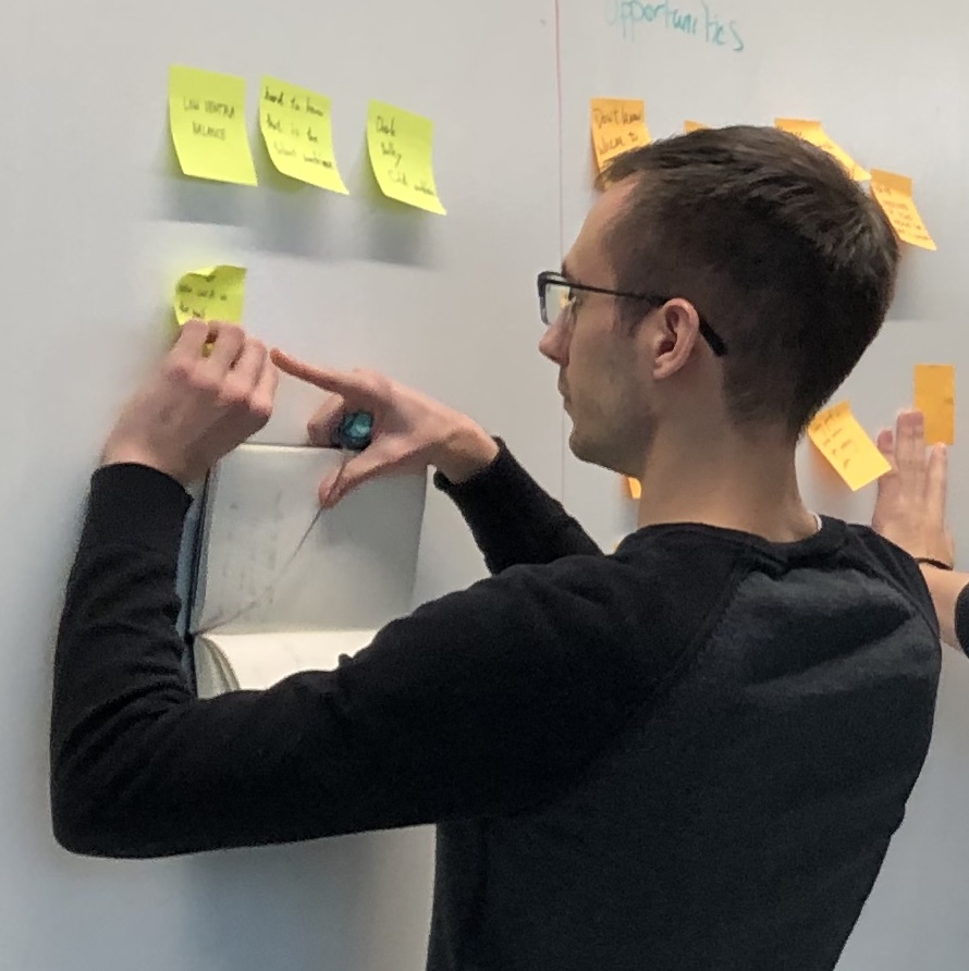 An image of Noah participating in a research synthesis workshop. He is holding a notebook and looking intently at a whiteboard as he adds a new sticky note to a group of ideas.